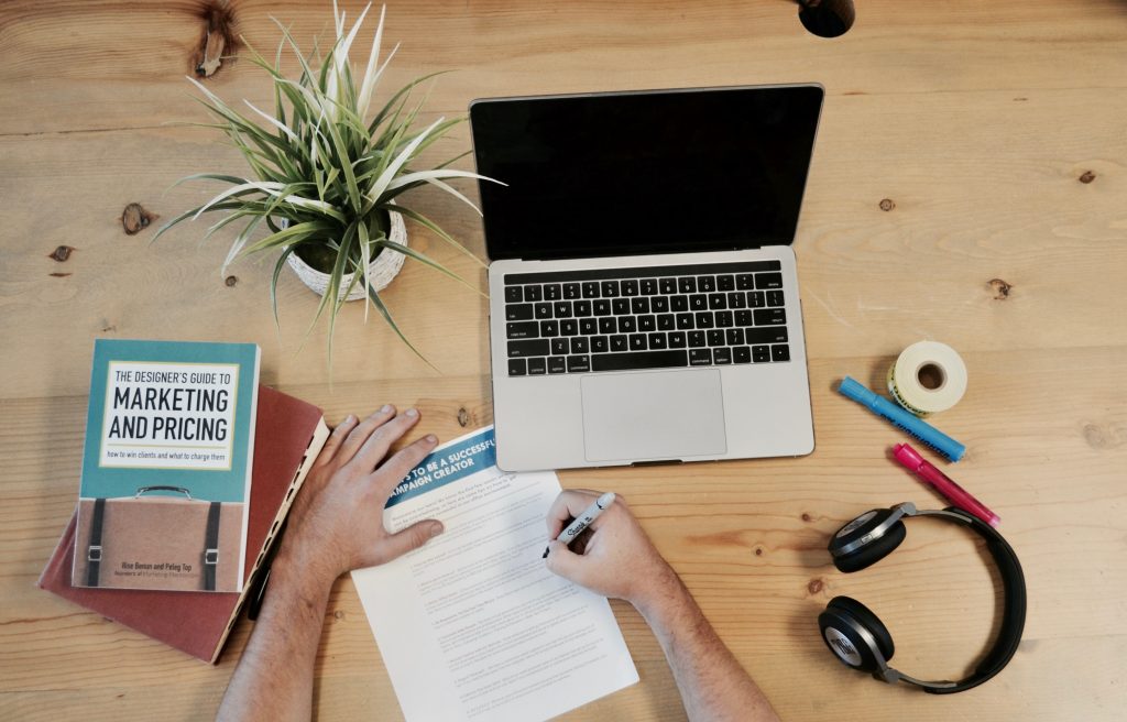 man writing in notebook next to laptop and headphones and plant